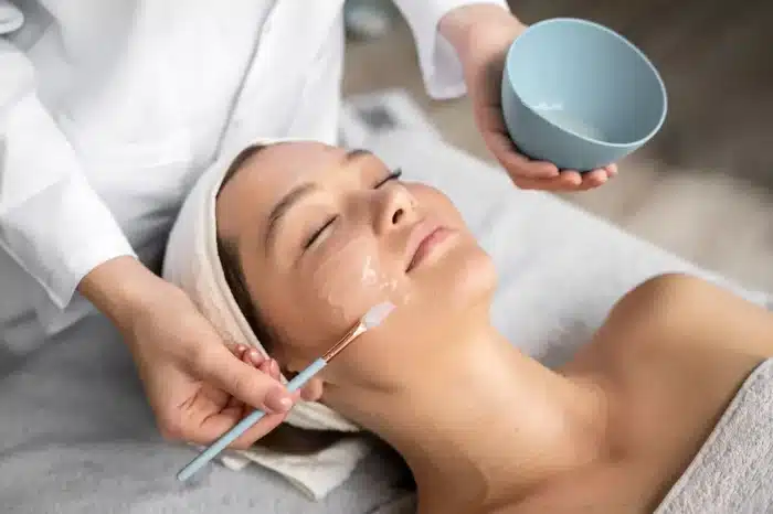 A person lies on a spa bed with eyes closed while a skincare specialist applies a facial mask using a brush. The person has a towel wrapped around their head and the specialist holds a small blue bowl.