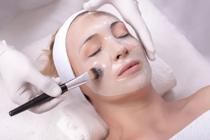 A woman with closed eyes lies on a spa bed, wearing a white headband and a clear facial mask. A gloved hand applies product to her face with a brush. The setting appears clean and relaxing.