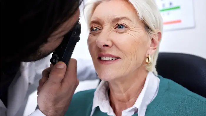 A doctor examines an older woman’s eye with a handheld device during an eye checkup. The woman, with short gray hair and gold hoop earrings, is smiling and seated in a clinic.
