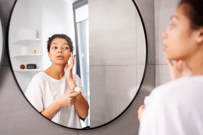 A woman in a white shirt applies PRX-T33 aftercare cream to her face while looking at herself in a round bathroom mirror. Shelves and a window are visible in the background.