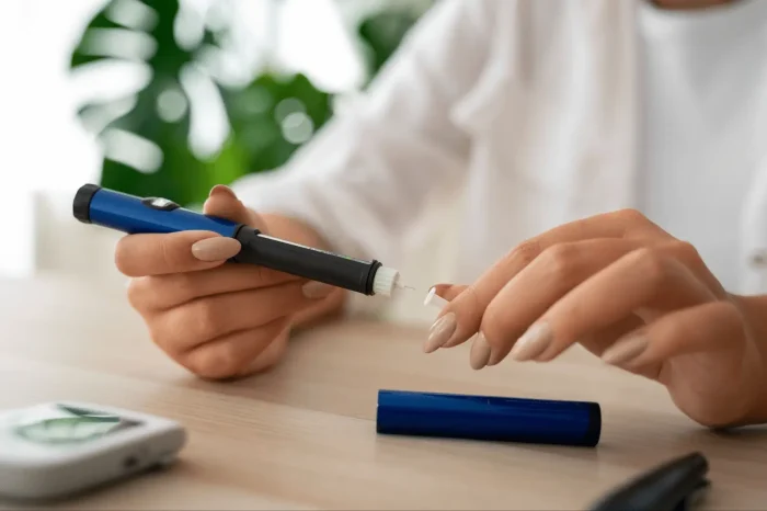 A person holds an insulin pen, preparing to use it, with a glucose meter and its case on a wooden table. The background is softly blurred with green plants visible.