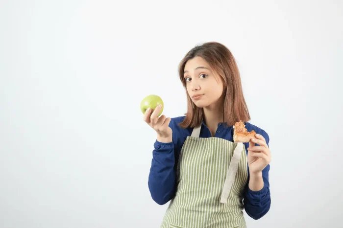 A young woman in an apron holds a green apple in one hand and a partially eaten pastry in the other, looking thoughtfully at the camera against a plain white background.