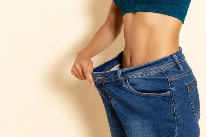 A person in a teal crop top shows weight loss by pulling the waistband of loose blue jeans away from their slim waist against a beige background.