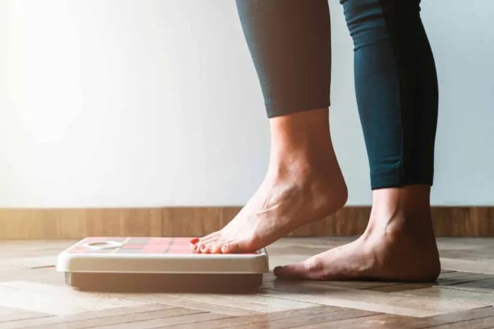 A person with bare feet and dark leggings stands on a digital bathroom scale on a wooden floor, checking their weight.