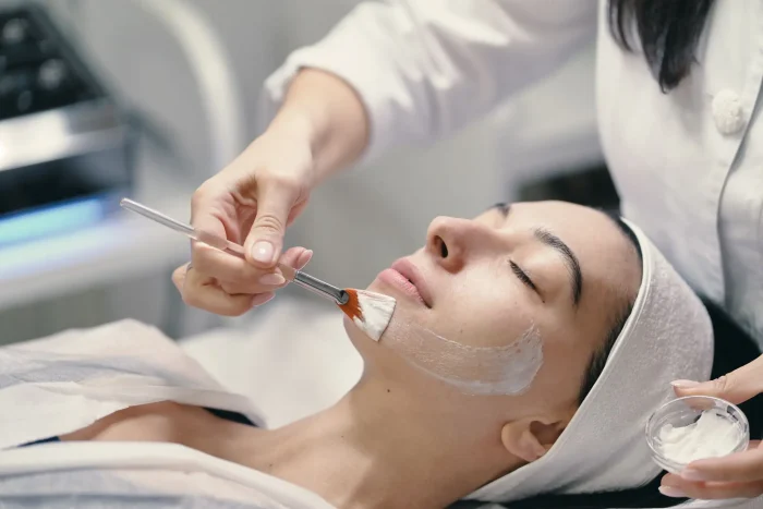 A person lying down with eyes closed receives a facial treatment as a practitioner applies a white cream mask to their face using a brush. The person wears a headband and a white robe.