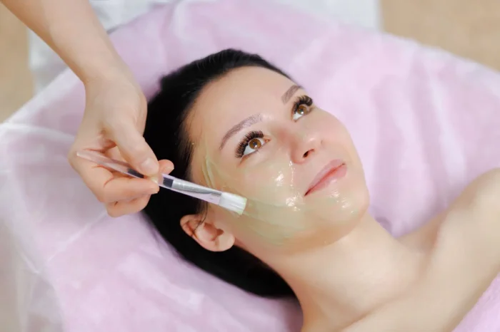 A woman lying down with her hair back receives a facial treatment as someone applies a transparent greenish face mask to her cheek with a brush. She looks relaxed and is covered with a pink towel.