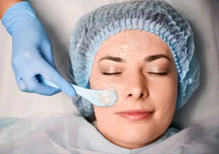 A person wearing a blue hair cap and protective gown lies with eyes closed while a gloved hand applies a gel facial mask to their face using a spatula.