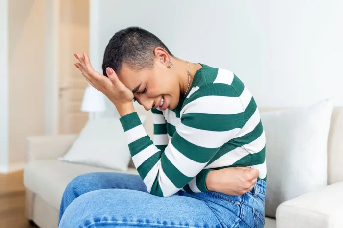 A person with short hair, wearing a green and white striped sweater and blue jeans, sits on a couch holding their stomach and forehead, appearing to be in pain or discomfort.