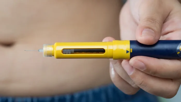 A person holds a yellow and blue injection pen with a visible needle, aimed towards their bare abdomen, suggesting preparation for a self-administered injection.