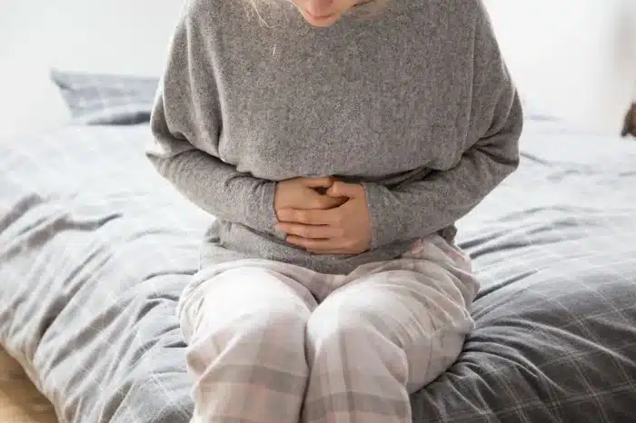 A person wearing a gray sweater and plaid pajama pants sits on a bed, holding their stomach with both hands, appearing to be in pain or discomfort.