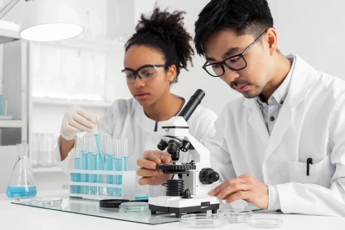 Two scientists in lab coats work in a laboratory; one examines samples under a microscope while the other handles test tubes filled with blue liquid. Both wear safety goggles, surrounded by lab equipment.