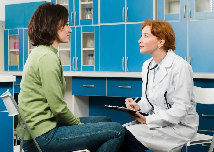 A doctor in a white coat with a stethoscope talks to a seated patient in a green sweater inside a medical office with blue cabinets. The doctor holds a clipboard and listens attentively.