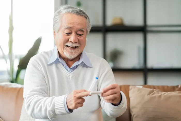 An older man with gray hair and a mustache, wearing a white sweater, sits on a couch and smiles while looking at and holding a small object in his hands in a bright, modern living room.