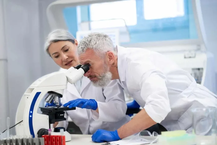 Two scientists in lab coats and blue gloves work together in a laboratory; one examines a sample through a microscope while the other observes, both surrounded by lab equipment.
