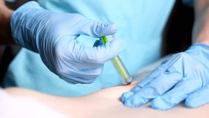 A healthcare professional wearing blue gloves administers an injection with a green syringe into a patient’s skin. The focus is on the hands and syringe, with a blurred background.