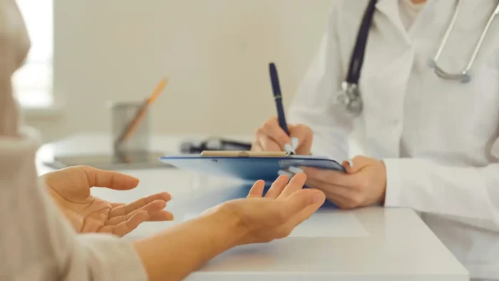 A doctor in a white coat writes on a clipboard while a patient sits across the desk, gesturing as they discuss Evenity side effects during the medical consultation.
