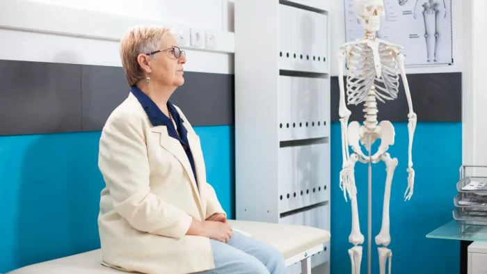 An older woman sits on a medical exam table in a doctor's office, looking at a human skeleton model displayed nearby. The room has blue and white walls with medical charts in the background.