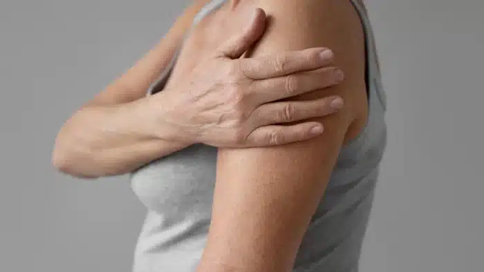 A person wearing a gray sleeveless top is touching their upper arm with their opposite hand, possibly indicating discomfort or pain in the arm or shoulder. The background is plain and neutral.