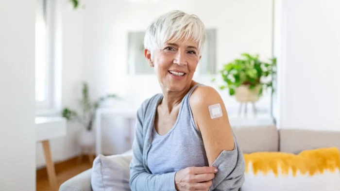 A smiling older woman with short white hair shows her upper arm with a bandage, sitting on a couch in a bright living room with plants in the background.