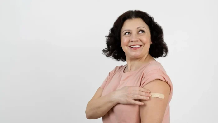 A smiling woman with short dark hair wearing a pink shirt shows her upper arm with a bandage, suggesting she recently received a vaccination. She is looking to the side against a plain white background.