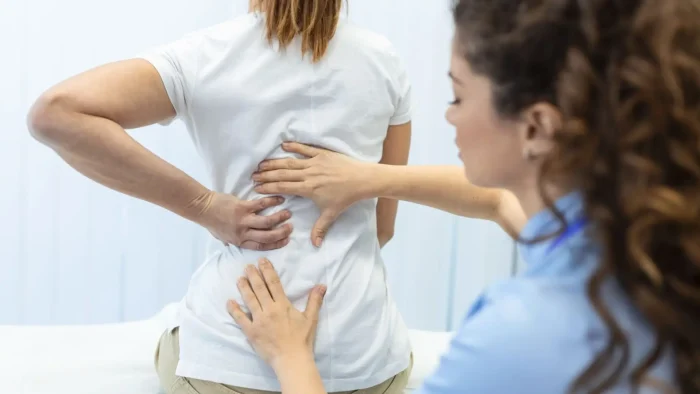 A healthcare professional examines a woman’s lower back as the woman touches her own back, indicating discomfort or pain during a consultation.