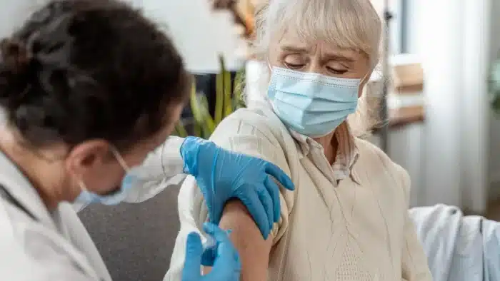 An elderly woman wearing a face mask receives a vaccine injection in her upper arm from a healthcare worker wearing gloves and a mask. The woman looks at her arm as the shot is administered.