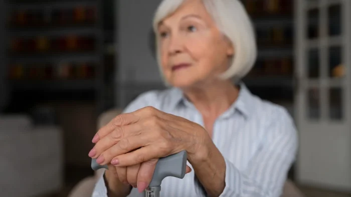 Elderly woman with short gray hair and a striped shirt sits indoors, resting her hands on top of a cane, looking thoughtfully into the distance. The background is softly blurred.